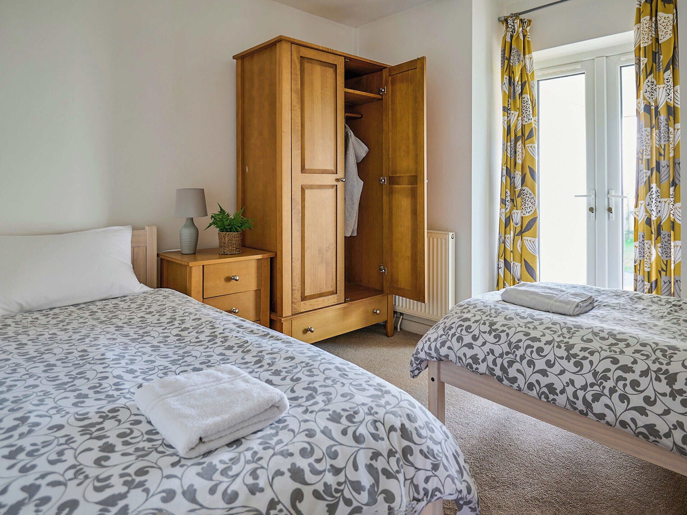 A bedroom with two beds, a wooden wardrobe, a bedside table with a lamp, and a window with curtains.