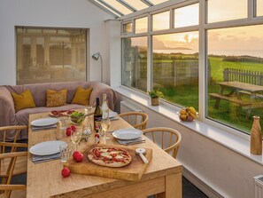 Un coin repas avec une table dressée pour deux personnes, un canapé avec des coussins, une vue sur un champ verdoyant à travers de grandes fenêtres et une table en bois.