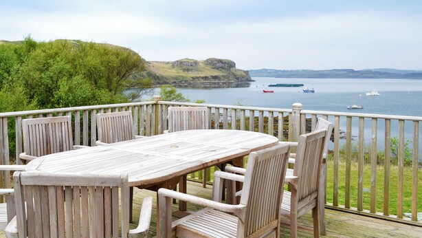 A wooden deck with a table and chairs overlooking a body of water with boats and a distant hill. - 4 bedroom accommodation in Portnalong, Isle of Skye (Portnalong, Isle of Skye)