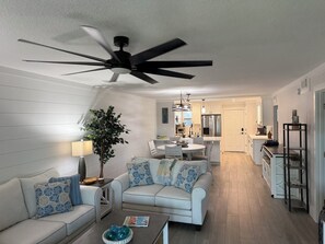 A modern living room with a ceiling fan, a dining area with a table and chairs, and a kitchen in the background.