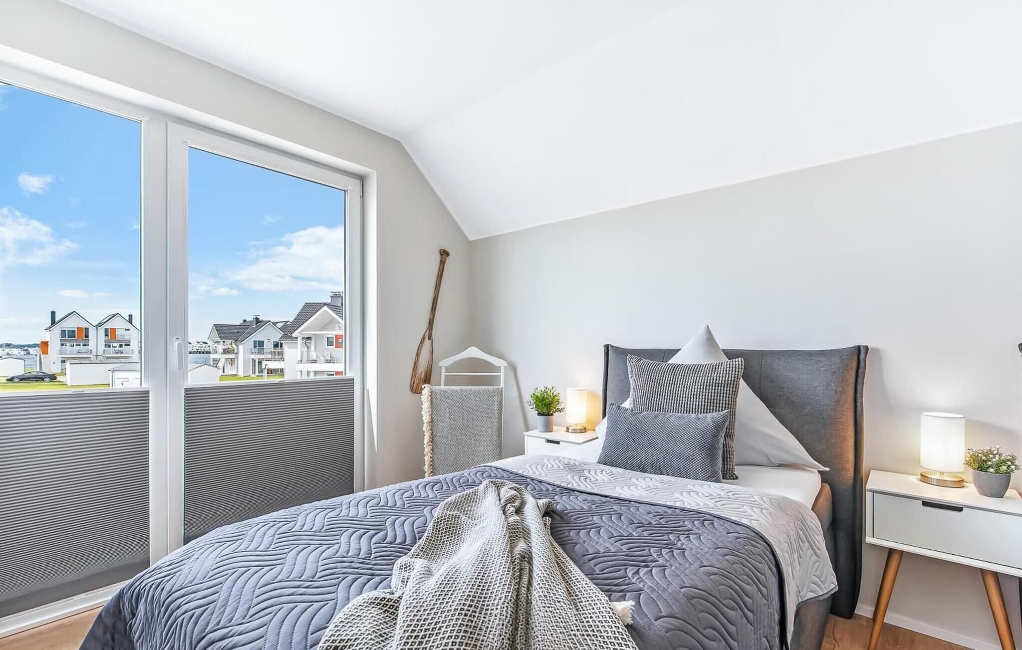 Un dormitorio con un ventanal grande, una cama con ropa de cama estampada, una mesita de noche con una lámpara y vistas a casas.