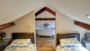 Un dormitorio con dos camas, techo de madera, un banco empotrado y un baño pequeño con lavabo y espejo.