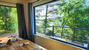 A wooden dining table with neatly folded napkins and a glass of water, set against a large window overlooking a lake and lush greenery.
