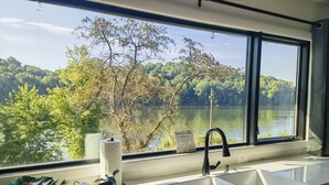 A kitchen with a large window overlooking a lake and trees. There is a roll of paper towels, a small decorative item, and a basket of packaged goods on the counter.