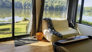 A room with a large window overlooking a lake, a bed, a small table, and a chair.