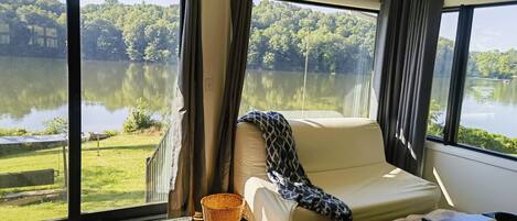 A room with a large window overlooking a lake, a bed, a small table, and a chair.