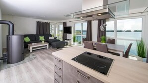 Uma sala de estar moderna com sofá, mesa de centro e área de jantar com mesa e cadeiras. Há uma cozinha com fogão e uma grande janela com vista para um corpo d'água.
