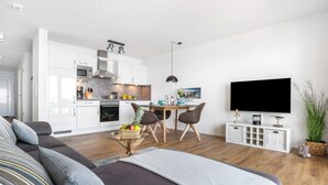 Una sala moderna con un sofá, una mesa de comedor con sillas, una zona de cocina con electrodomésticos y un televisor montado en la pared.