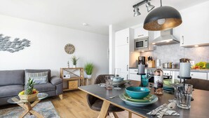 Una sala de estar moderna con un sofá gris, una mesa de comedor con platos, vasos y una botella de vino. Se ve una cocina con electrodomésticos modernos al fondo.