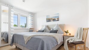 Un dormitorio con cama, una silla, una mesita de noche, un ventanal y un cuadro en la pared.
