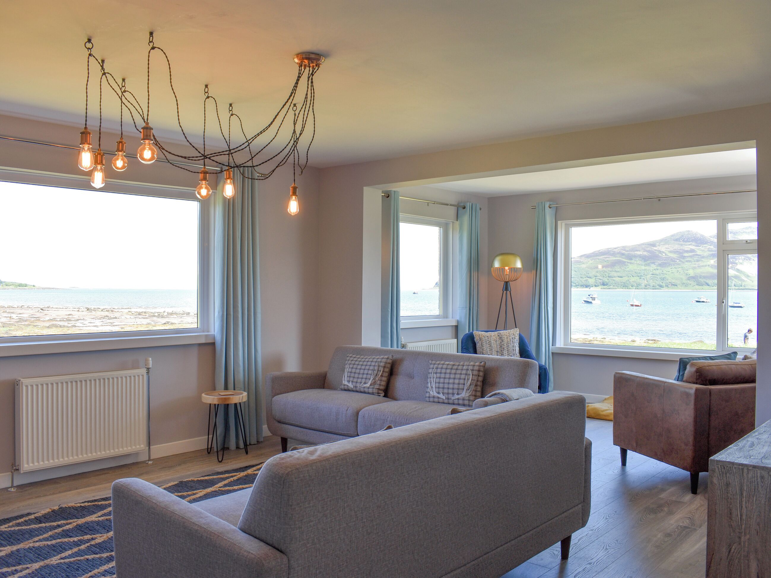 A living room with a sofa, armchairs, and a floor lamp. There are windows with curtains and a view of the sea.