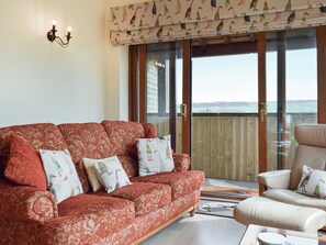 A red patterned sofa with pillows, a white armchair, and a glass door leading to a balcony with a view. - 4 bedroom accommodation in Holmfirth (Holmfirth)