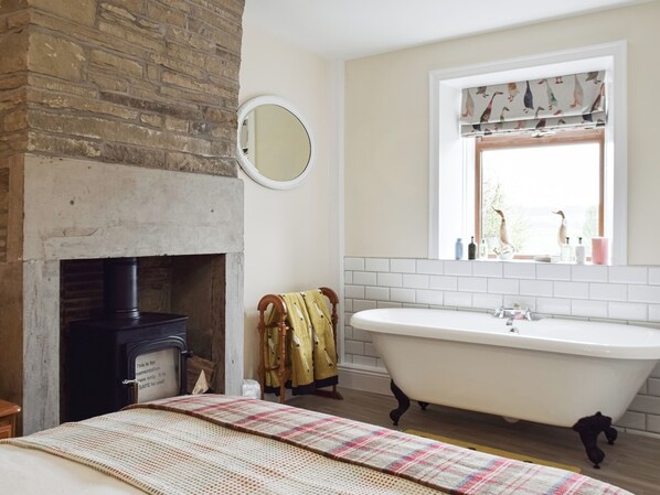 Un baño con una bañera independiente, un espejo, una chimenea y un ventanal con cortinas estampadas.