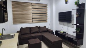 A modern living room with a sectional sofa, a small ottoman, a TV on a stand, and a kitchen area with a sink. - SHEPHERD LINKS APARTMENTS  (Accra)