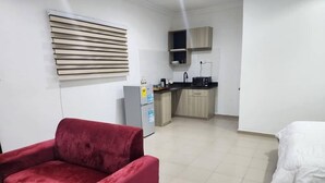 A compact room with a red armchair, a bed with white bedding, a kitchenette with a microwave, and a small cabinet. - SHEPHERD LINKS APARTMENTS  (Accra)