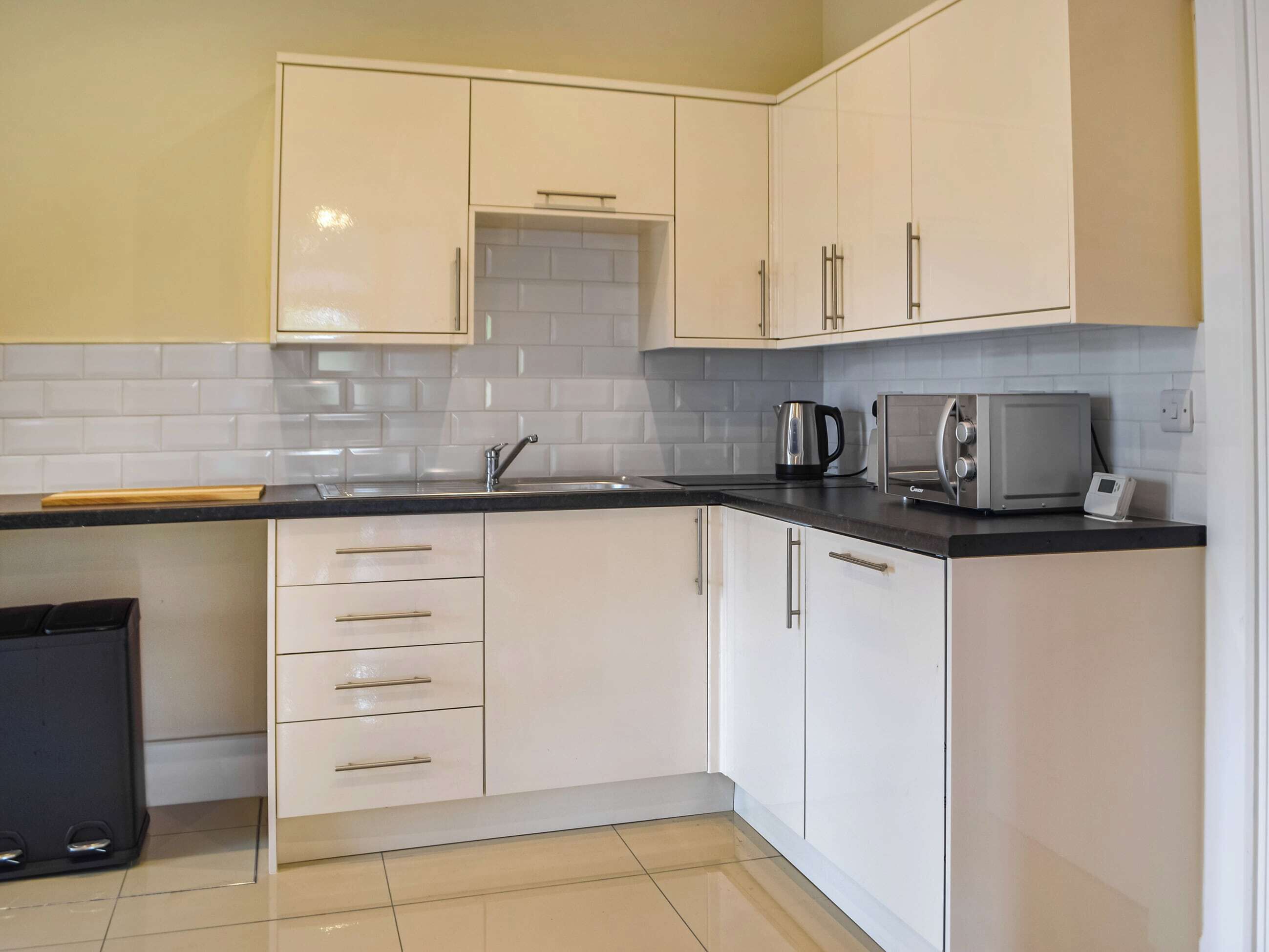 A kitchen with white cabinets, a black countertop, a kettle, a microwave, and a trash bin.