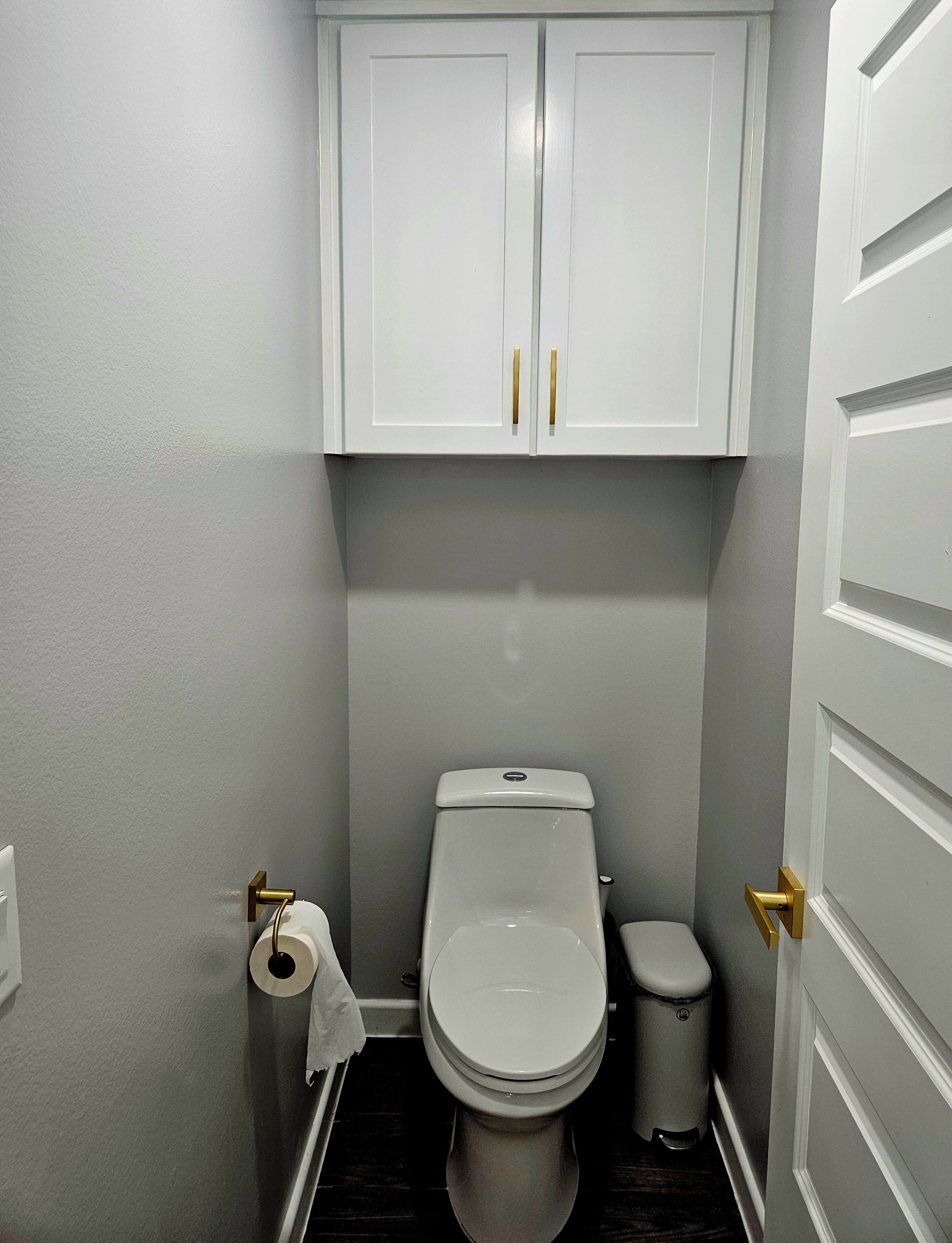 A small bathroom with a toilet, a trash can, and a roll of toilet paper on the floor.