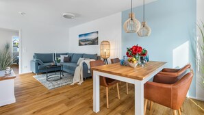 A modern living room with a blue sofa, a wooden dining table with four chairs, and a large window.