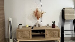 A modern living room with a flat-screen TV mounted on the wall, a wooden entertainment unit with decorative items, and a standing fan. - Appartement les Clés de Juliette (Gruissan)