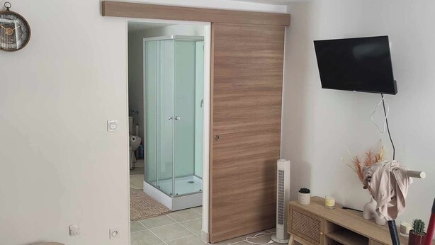 A modern bathroom with a glass shower enclosure, a wooden vanity, and a mounted TV. - Appartement les Clés de Juliette (Gruissan)