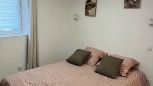A bedroom with a checkered bedspread, two pillows, a window with blinds, and two wall-mounted artworks. - Appartement les Clés de Juliette (Gruissan)