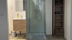 A modern bathroom with a glass-enclosed shower, a vanity with a mirror, and a wooden shelving unit. - Appartement les Clés de Juliette (Gruissan)