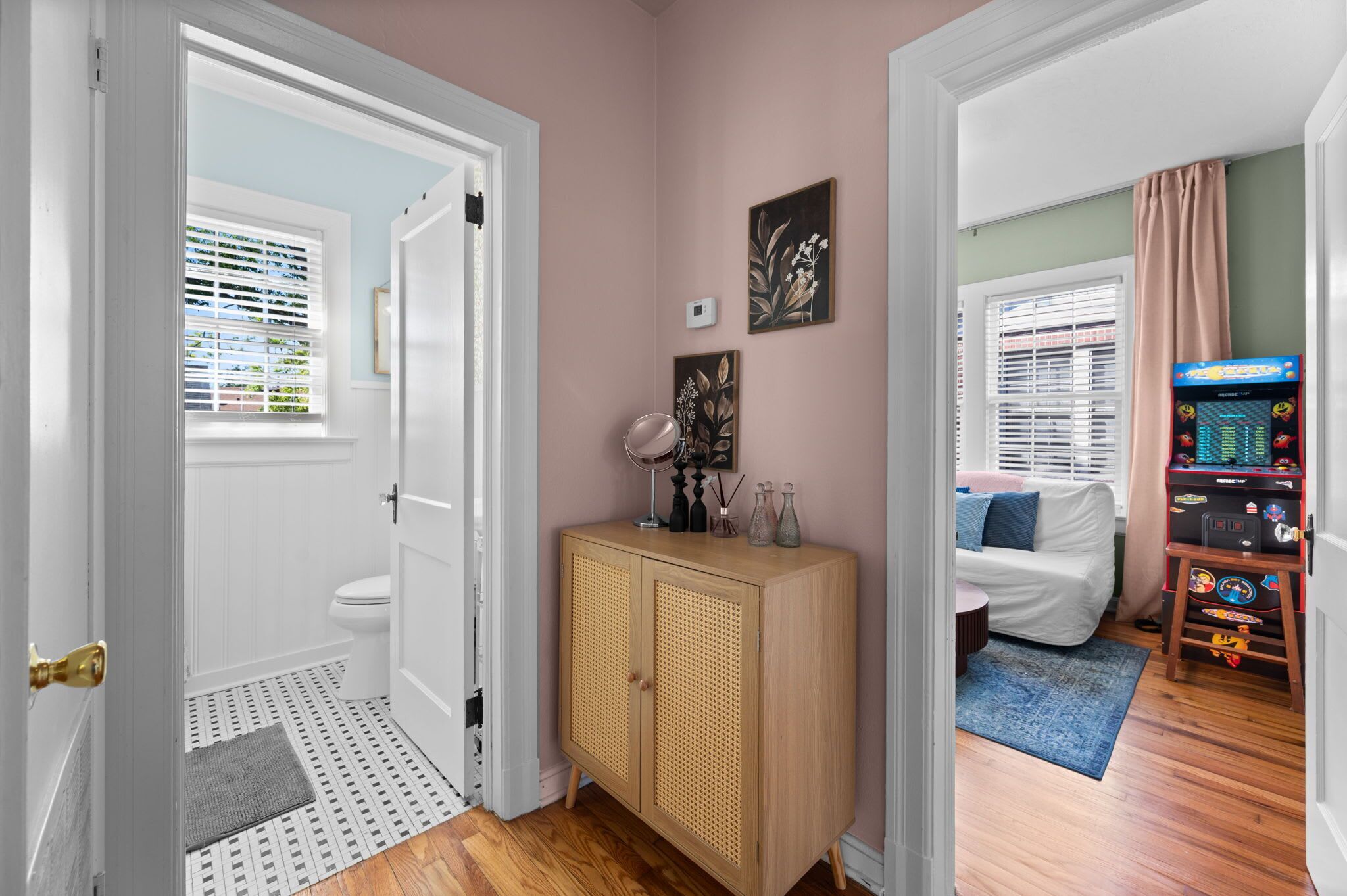 A hallway with a door leading to a bathroom, a wooden cabinet with decorative items, and a room with a bed and a gaming console.
