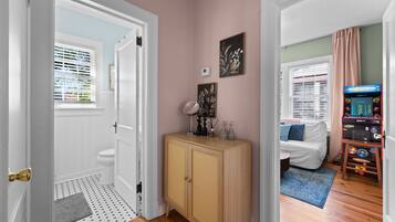A hallway with a door leading to a bathroom, a wooden cabinet with decorative items, and a room with a bed and a gaming console.