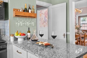A kitchen with a granite countertop, wine bottles, glasses, and a fruit bowl.