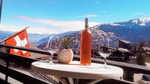 Una mesa con una botella, una piña y dos copas de vino en un balcón con una bandera suiza y vistas a la montaña.