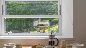 Une table en bois, près d’une fenêtre donnant sur un paysage verdoyant, est dressée pour le petit-déjeuner : on y trouve des bols étiquetés pour les céréales, la soupe, le café et le thé, une cafetière et des croissants.