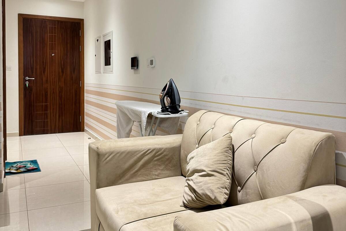 A modern living room with a beige sofa, a small table with an iron, and a door in the background.