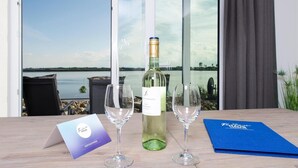 A table with a bottle of wine, two glasses, a menu, and a view of a lake through large windows. - Yachtblick auf der Ostsee (Olpenitz)