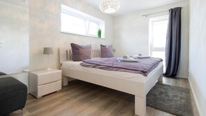 A modern bedroom with a white bed, two bedside tables, a grey sofa, a window with curtains, and a hanging light fixture. - Yachtblick auf der Ostsee (Olpenitz)