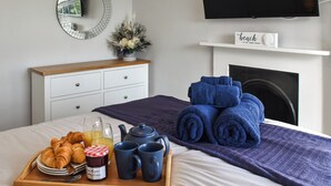 Un dormitorio ordenado con cama, una mesita de noche con alimentos para el desayuno, un espejo, una chimenea y un televisor.