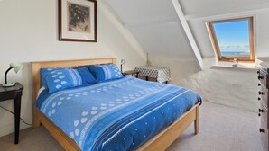 Una camera da letto con soffitto spiovente, un letto con biancheria azzurra, un comodino con una lampada, un tavolino con tovaglia decorata e una finestra vista mare.