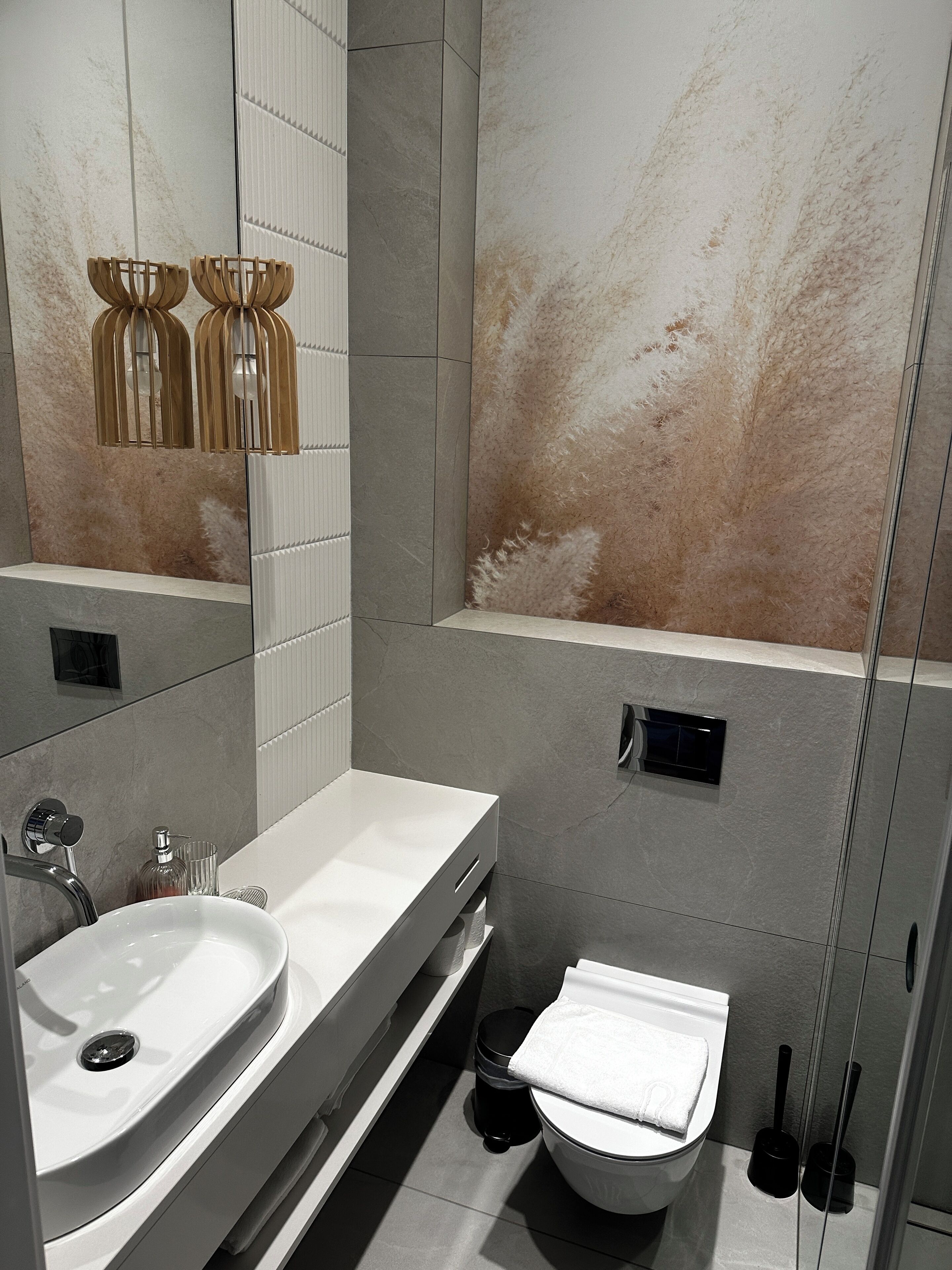A modern bathroom with a white sink, mirror, and toilet. There are two black trash bins and a towel rack.