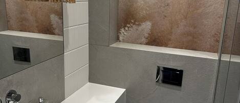 A modern bathroom with a white sink, mirror, and toilet. There are two black trash bins and a towel rack.