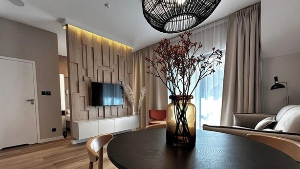 A modern living room with a round dining table, a flat-screen TV mounted on a wooden wall, and a large window with curtains. - Gościniec Warmiński by Elite4Rent (Stawiguda)