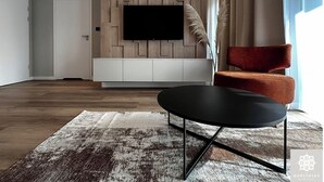 A modern living room with a flat-screen TV mounted on a wooden wall, a round black coffee table, a red armchair, and a patterned rug. - Gościniec Warmiński by Elite4Rent (Stawiguda)