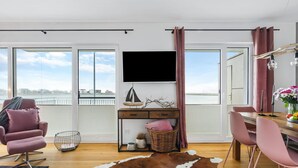 Una sala moderna con un ventanal grande, un televisor de pantalla plana, una mesita de madera y una zona de comedor con sillas rosadas.