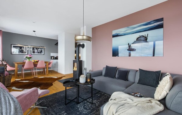 Sala moderna com sofá cinza, mesa de centro e lareira. Há um espaço de jantar com cadeiras cor-de-rosa e mesa de madeira. Um quadro grande está na parede, e o ambiente apresenta um design moderno com um tapete preto.