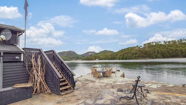 A dock with a staircase leading to a building, a table and chairs by the water, and a view of a forested hill.