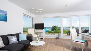 Una sala de estar moderna con un televisor grande, un sofá, una mesita y una silla. Hay un balcón con vistas al mar.