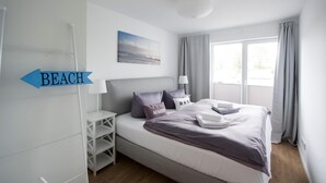 Un dormitorio con cama, almohadas, una mesita de noche, lámpara, ventana con cortinas y un cartel que indica el camino a la playa.