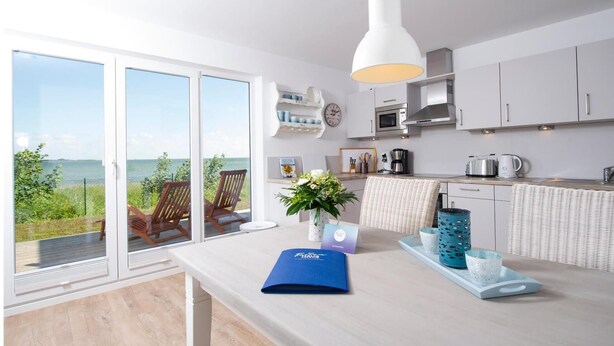 Una cocina moderna con vista al mar, armarios blancos y un comedor con mesa y sillas.