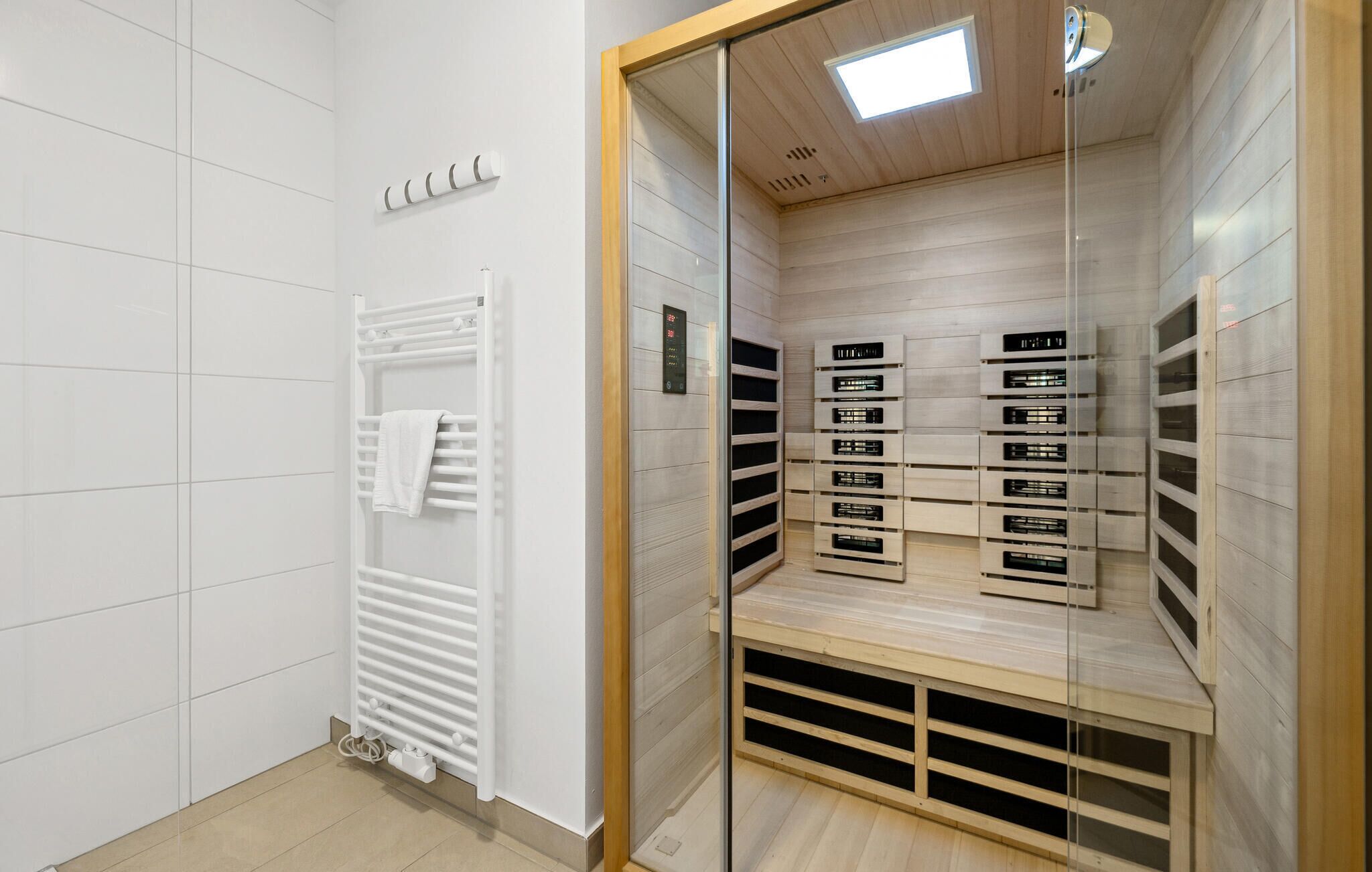 A modern bathroom with a white tiled wall, a built-in towel rack, a glass-enclosed shower area with a wooden bench, and a visible control panel.