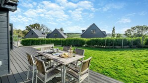 A wooden deck with a table set for dining, surrounded by chairs, and a view of a green lawn and houses in the background. - Fair Isle Holiday Cottages - Fisher (Near Newport)