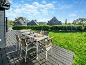 A wooden deck with a table set for dining, surrounded by chairs, and a view of a green lawn and houses in the background. - Fair Isle Holiday Cottages - Fisher (Near Newport)