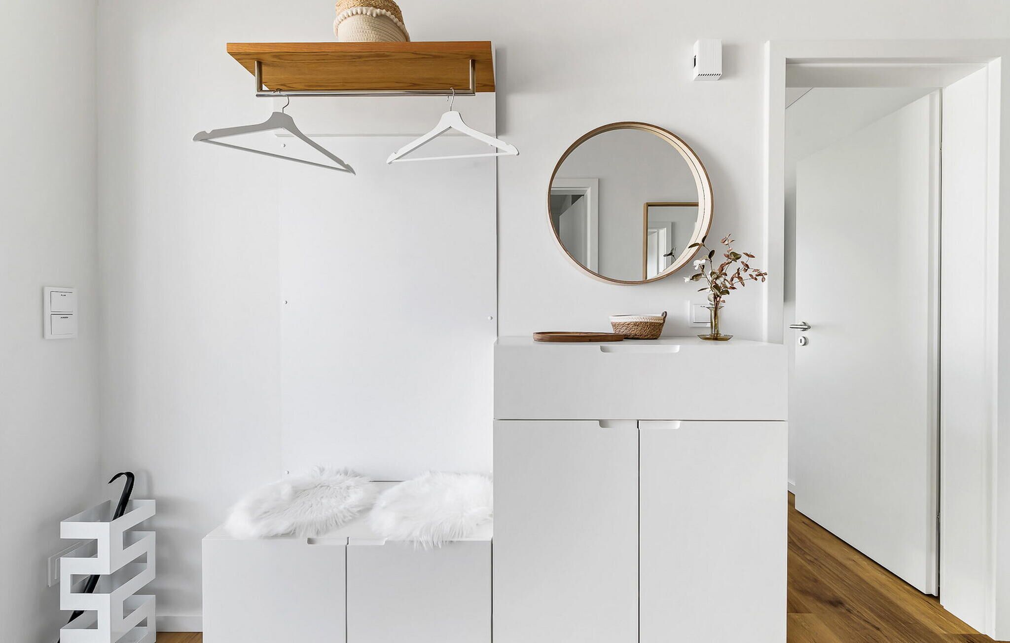 Une salle de bain minimaliste avec une étagère en bois, deux cintres blancs, un miroir rond et un meuble-lavabo blanc.